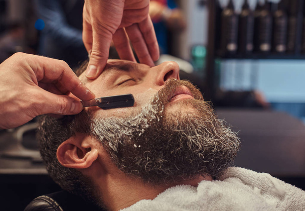 Szép csípő szakállas férfi ül a karosszékben, egy fodrász boltban, míg a fodrász leborotválja a szakállát egy veszélyes borotva. - Fotó, kép