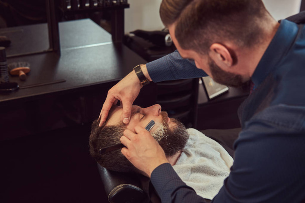 Bebaarde mannen zitten in een leunstoel in een kapper terwijl kapper zijn baard met een gevaarlijke scheermes scheert. - Foto, afbeelding