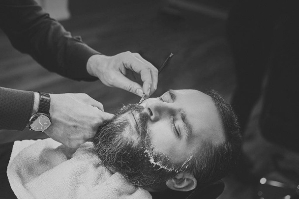 A handsome hipster bearded male sitting in an armchair in a barber shop while hairdresser shaves his beard with a dangerous razor. - Photo, Image