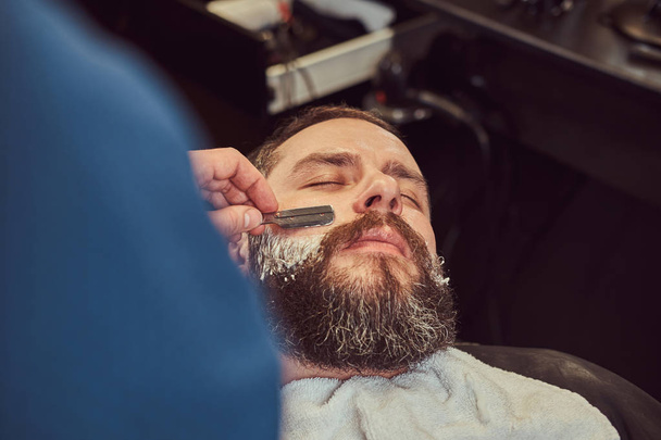 Красивый бородатый хипстер сидит в кресле в парикмахерской, пока парикмахер бреет бороду опасной бритвой
. - Фото, изображение