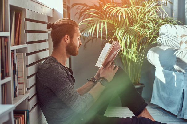Retrato de un guapo estudiante pelirrojo con ropa casual, sentado en un piso en una casa mientras lee un libro, mirando a una cámara
. - Foto, Imagen