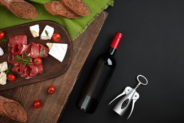 Wine bottle with cheese and traditional sausages on wooden board on black background with copy space - Фото, зображення