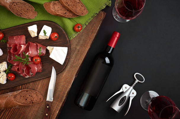 Red wine, cheese, cherry tomato, bread and prosciutto on wooden board over black backdrop, top view, copy space - Photo, Image