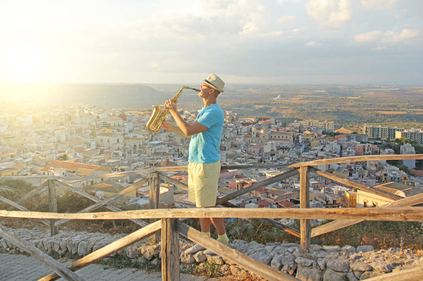 Un hombre con sombrero toca el saxofón en la naturaleza, sobre el fondo de una puesta de sol. El hombre es músico. La isla de Sicilia, Italia
. - Foto, Imagen