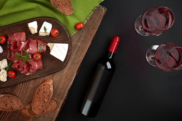 Red wine, cheese, cherry tomato, bread and prosciutto on wooden board over black backdrop, top view, copy space - Fotoğraf, Görsel