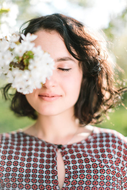 weather, lifestyle, tenderness concept. there is portrait of young and happy girl with delicate smile and closed eyes, blooming flowers of apple tree are covered her face - Photo, image