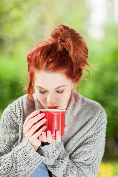beautiful young woman with red hair sitting in the garden, relaxing, drinking coffee in a red coffee mug - Fotografie, Obrázek