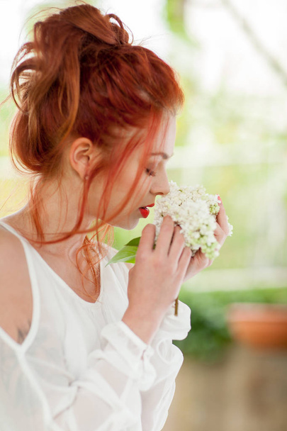 beautiful woman with red hair holding white lilac bloom in her hands, smelling fragrant - Фото, изображение