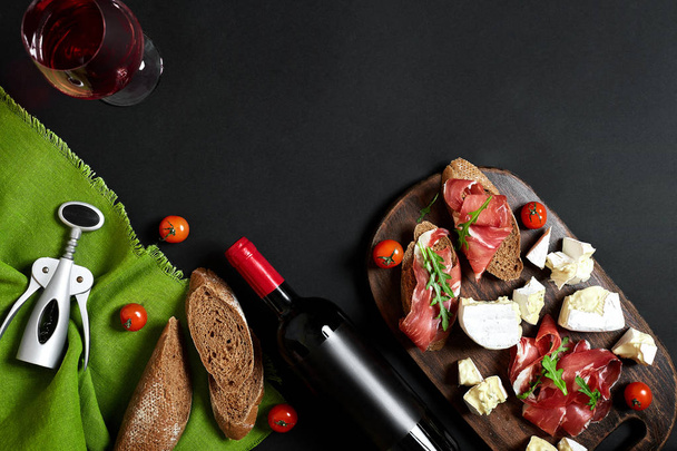 Delicious appetizer to wine - ham, cheese, baguette slices, tomatoes, served on a wooden board, and glass with red wine on black surface - Photo, Image