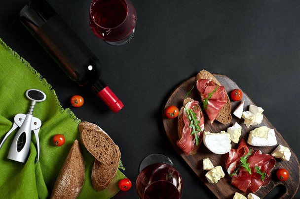 Κόκκινο κρασί, τυρί, ντομάτα, ψωμί και προσούτο σε ξύλινα του σκάφους πάνω από το μαύρο φόντο, το top view, αντίγραφο χώρου - Φωτογραφία, εικόνα