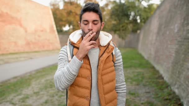 Молодой красивый мужчина наслаждается сигаретой в общественном парке
. - Кадры, видео