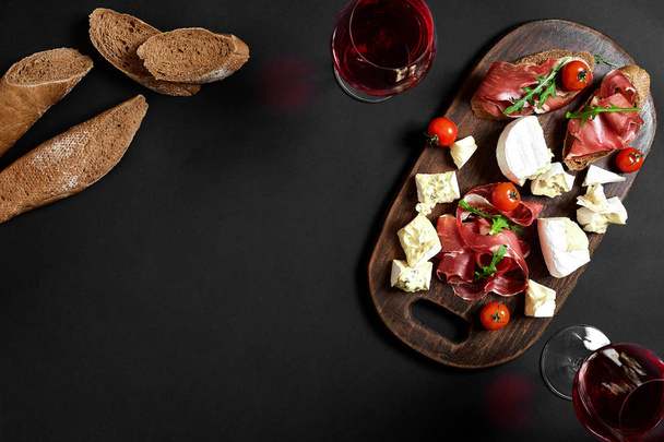 Delikátní předkrm k vínu - šunka, sýr, plátky bagety, rajčaty, podávané na dřevěném prkénku a skla s červeným vínem na černém povrchu - Fotografie, Obrázek