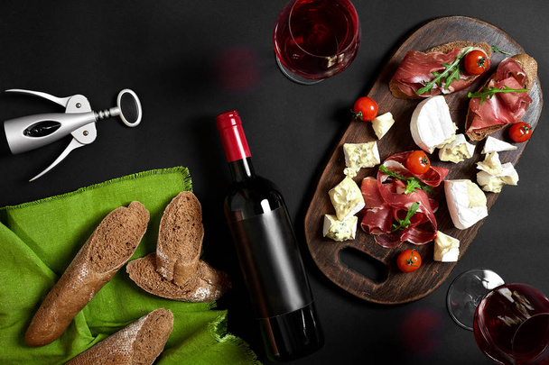 Νόστιμο ορεκτικό στο κρασί - ζαμπόν, τυρί, φέτες μπαγκέτα ντομάτες, σερβίρεται σε μια ξύλινη σανίδα, και γυαλί με το κόκκινο κρασί στην μαύρη επιφάνεια - Φωτογραφία, εικόνα