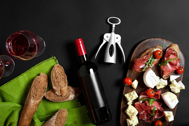Delicious appetizer to wine - ham, cheese, baguette slices, tomatoes, served on a wooden board, and glass with red wine on black surface - Photo, image