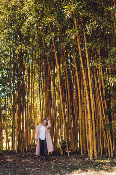 curly woman in a pink coat is standing next to tall bamboo trees, posing and resting - Foto, Bild