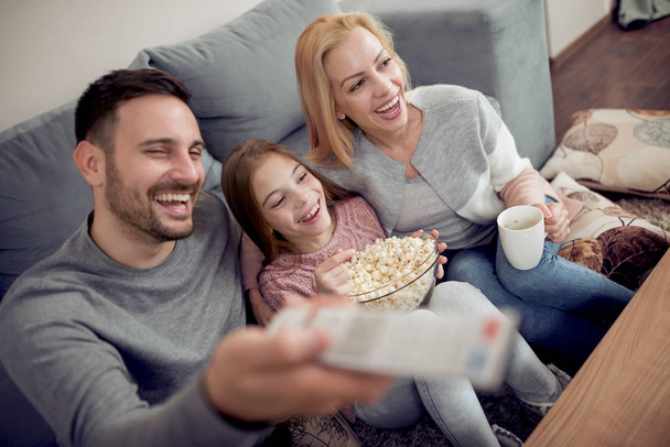 Πατέρα, μητέρα και κόρη, βλέποντας τηλεόραση και τρώγοντας ποπ κορν στο σαλόνι στο σπίτι. - Φωτογραφία, εικόνα