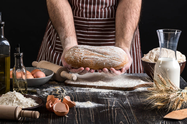 Кухар тримає свіжий хліб в руці. Чоловік готує тісто за столом на кухні
. - Фото, зображення