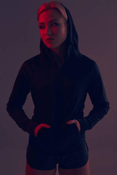 Mooie verleidelijke sportieve meisje met de slanke sexy figuur in de zwarte hoodie, sport ondergoed en kleine zwarte shorts is poseren in de studio - Foto, afbeelding