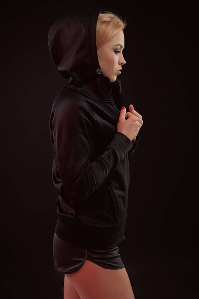 Belle fille sportive séduisante avec la silhouette sexy mince dans le sweat à capuche noir, sous-vêtements de sport et petit short noir pose en studio
 - Photo, image