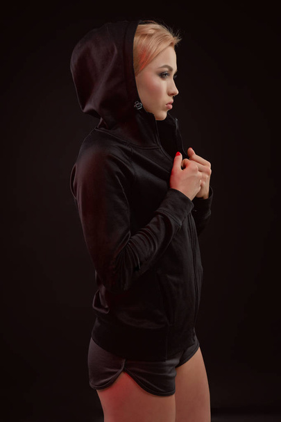 Beautiful seductive sporty girl with the slim sexy figure in the black hoodie, sport underwear and little black shorts is posing in the studio - Zdjęcie, obraz