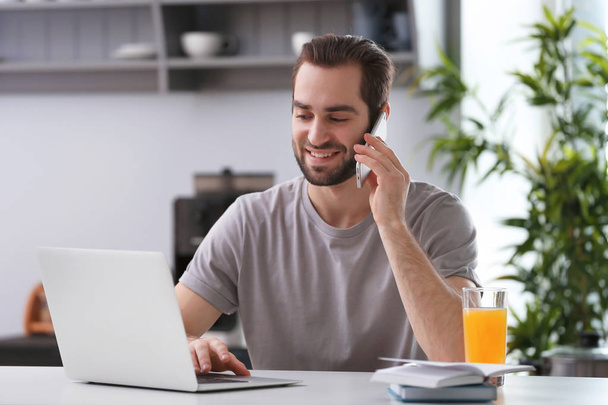 Αρσενικό freelancer μιλάμε στο τηλέφωνο ενώ εργάζεστε με το φορητό υπολογιστή στο γραφείο στο σπίτι - Φωτογραφία, εικόνα