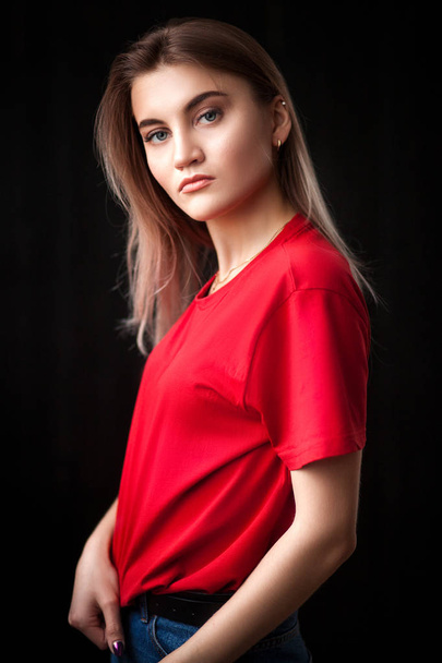Красивая молодая блондинка в красной футболке и джинсах улыбается и позирует на черном изолированном фоне
 - Фото, изображение