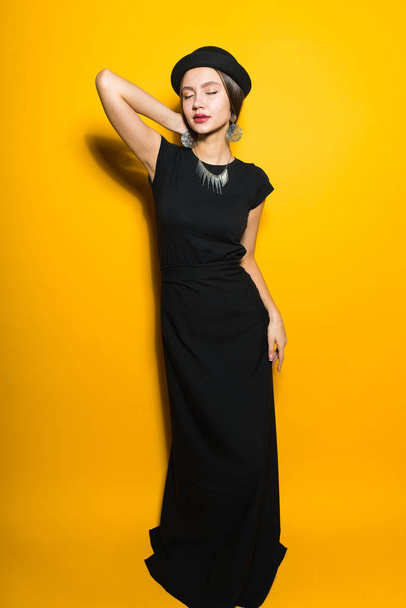 confident stylish girl model in a long black dress and hat posing on a yellow background - Φωτογραφία, εικόνα