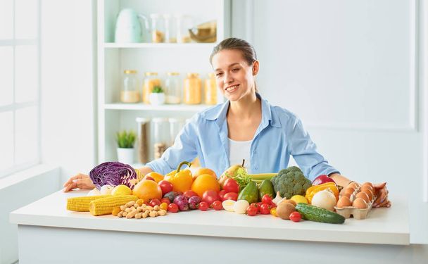 glückliche junge Hausfrau, die in der Küche sitzt und aus einem Haufen frischem Bio-Obst und -Gemüse Essen zubereitet - Foto, Bild