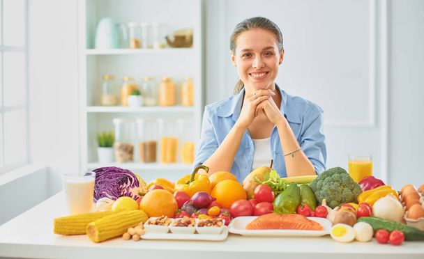 feliz jovem dona de casa sentada na cozinha preparando alimentos de uma pilha de frutas e legumes orgânicos frescos diversos
 - Foto, Imagem