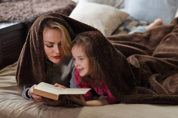 Mother and daughter lie on a bed under a blanket and read a book. - 写真・画像