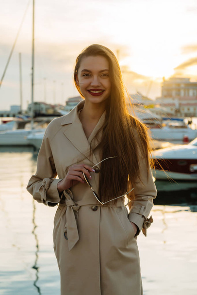 long-haired gorgeous girl in a trendy coat holds glasses in her hands, smiling at the seaport - Photo, image