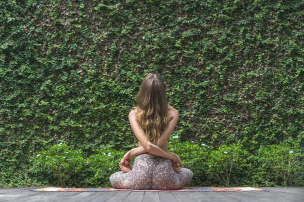 vista trasera de la mujer practicando yoga en pose de loto frente a la pared cubierta de hojas verdes
 - Foto, imagen