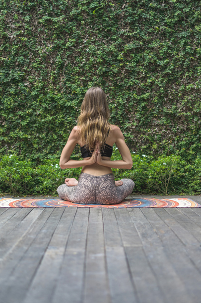 vista trasera de la mujer practicando yoga en pose de loto haciendo gesto namaste detrás de la espalda frente a la pared cubierta de hojas verdes
 - Foto, Imagen