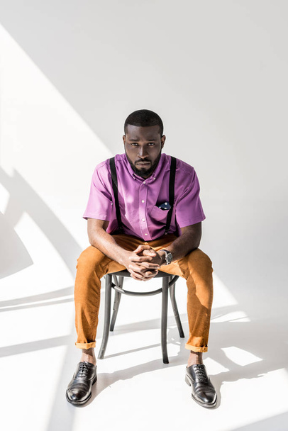 young african american man in stylish clothing with hands in lock sitting on chair on grey background - Photo, Image