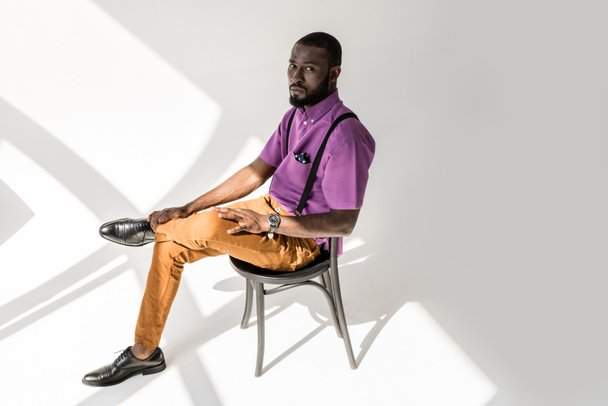 vista lateral del hombre afroamericano reflexivo en ropa de moda sentado en la silla sobre fondo gris
 - Foto, Imagen