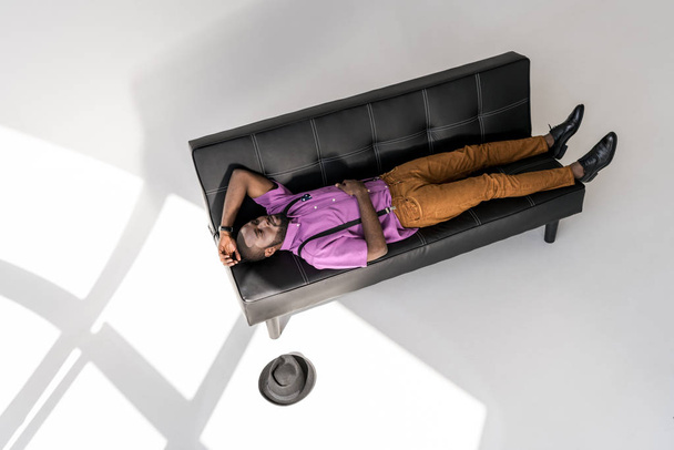 high angle view of african american fashionable man lying on black sofa with hat on floor - Photo, Image