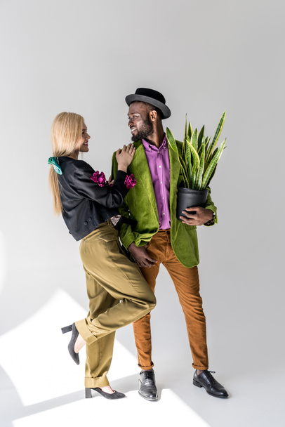 улыбающаяся межрасовая стильная пара с зеленым растением в цветочном горшке позирует на сером фоне
 - Фото, изображение