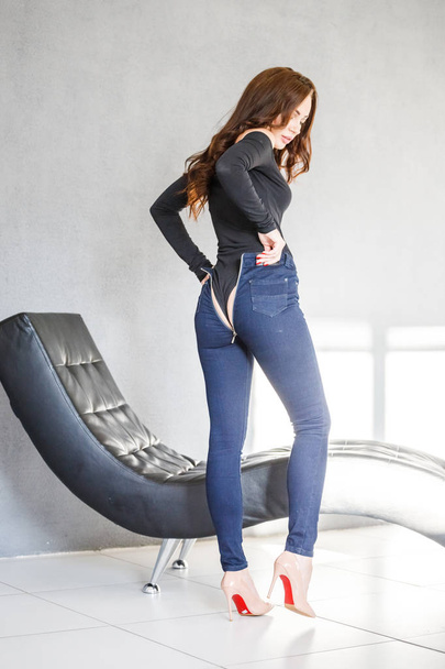 Studio portrait of beautiful brunette girl in blue jeans and black underwear standing close to dark sofa on white wall background - Φωτογραφία, εικόνα