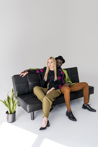 πολυπολιτισμική μοντέρνο ζευγάρι αναπαύεται στον μαύρο καναπέ σε γκρι φόντο - Φωτογραφία, εικόνα
