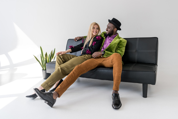 fröhliches multikulturelles Modepaar auf schwarzem Sofa auf grauem Hintergrund - Foto, Bild