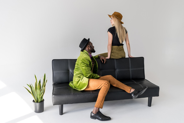мультикультурная модная пара в шляпах на черном диване на сером фоне
 - Фото, изображение
