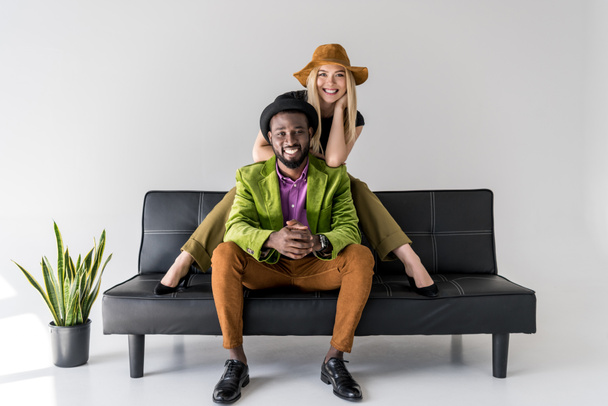 веселая мультикультурная модная пара в шляпах, сидящая на черном диване на сером фоне
 - Фото, изображение