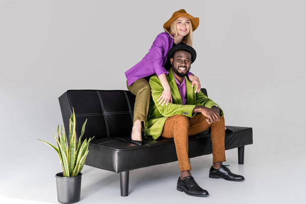 cheerful multicultural fashionable couple in hats sitting on black sofa on grey background - Photo, Image