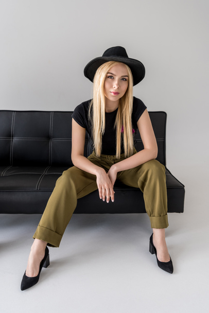 attractive woman in  hat sitting on black sofa on grey backdrop - Fotografie, Obrázek