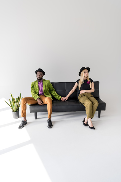 young multiethnic couple holding hands while sitting on couch and looking away on grey  - Fotó, kép
