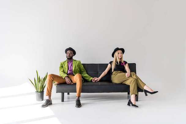 stylish young multiethnic couple holding hands while sitting on sofa and looking away on grey  - Foto, Imagem