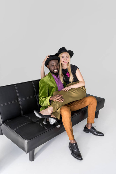 high angle view of fashionable multiethnic couple in hats sitting on sofa and smiling at camera on grey - Zdjęcie, obraz