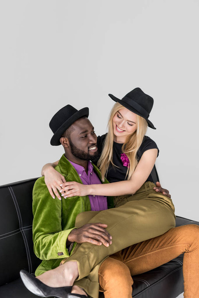 bonito elegante jovem casal multiétnico abraçando e sorrindo no sofá isolado em cinza
 - Foto, Imagem