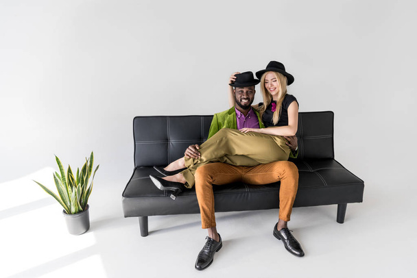 happy young multiethnic couple in stylish clothes sitting on sofa and smiling at camera on grey   - Fotografie, Obrázek