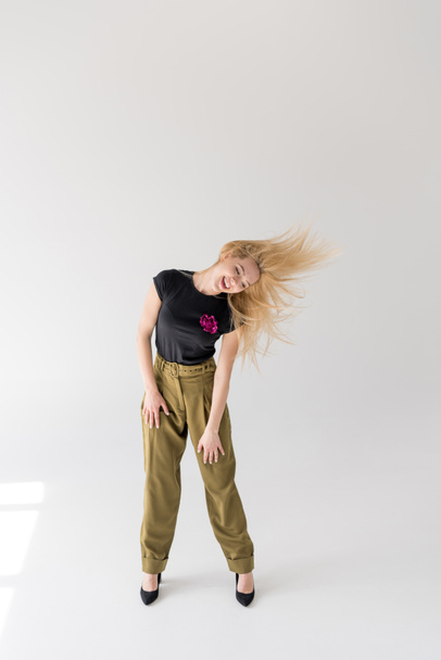full length view of cheerful blonde girl dancing and shaking hair on grey - Foto, imagen